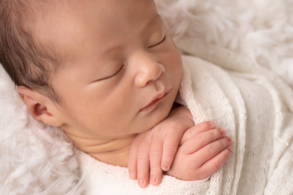 baby close up face and tiny hands tucked under his chin wrapped in white in white fur