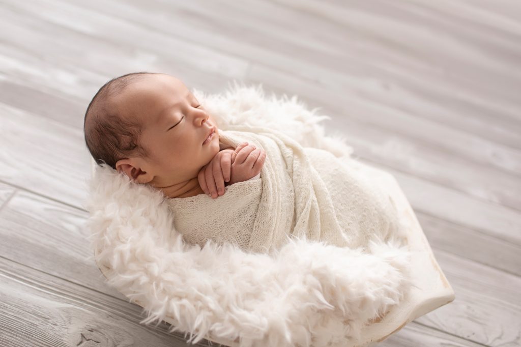 baby wrapped in white against back light in white fur stuffed bowl against grey floor