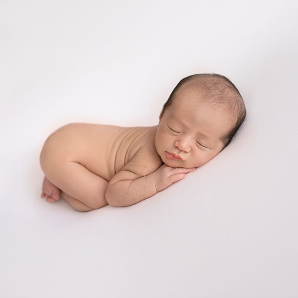 newborn naked posed on his belly on white blanket head resting on his hands bottoms up face to camera