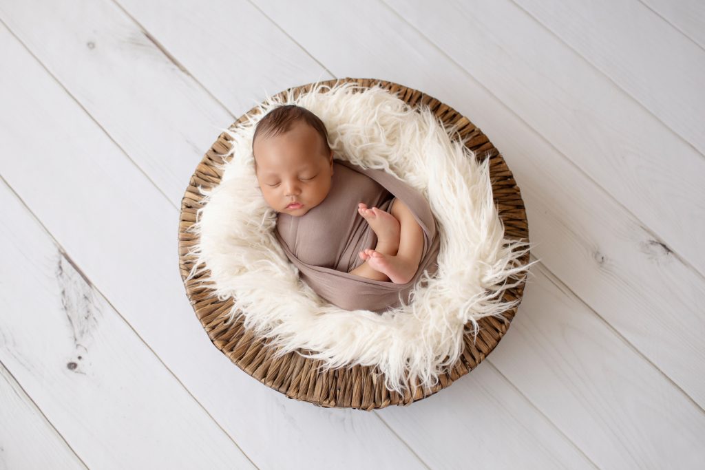 Lucas wrapped in brown swaddle in white fur stuffed wicker basket on white wood floor