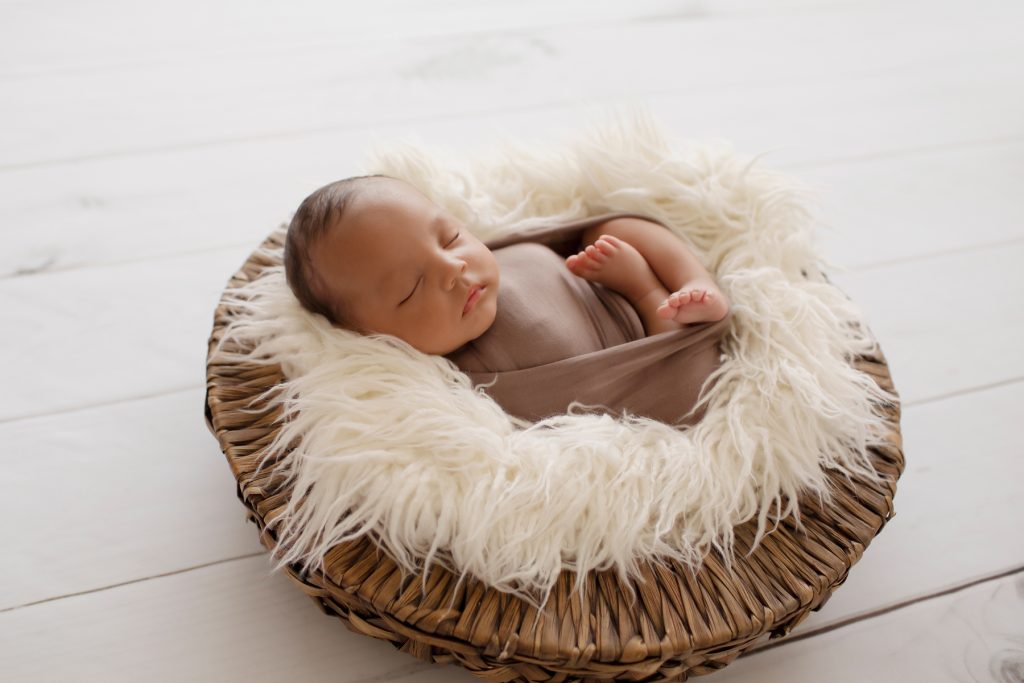 Lucas wrapped in brown swaddle in white fur stuffed wicker basket on white wood floor with backlight