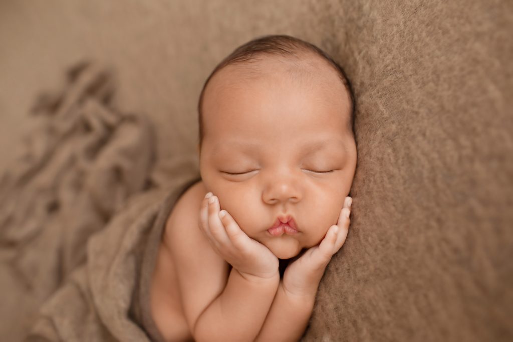 Lucas on brown blanket with little baby hands cupping his beautiful baby face toward camera