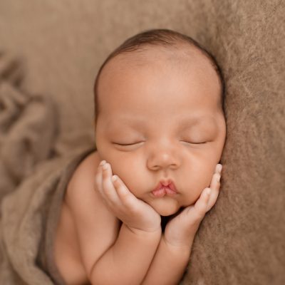 Lucas on brown blanket with little baby hands cupping his beautiful baby face toward camera