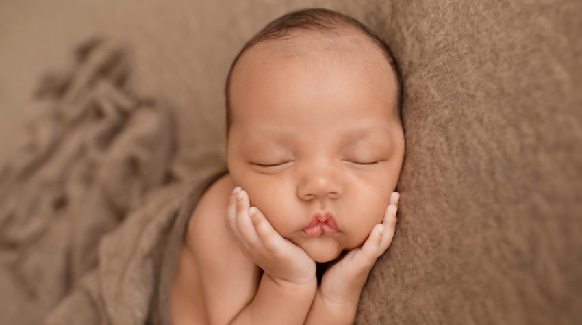 Lucas on brown blanket with little baby hands cupping his beautiful baby face toward camera