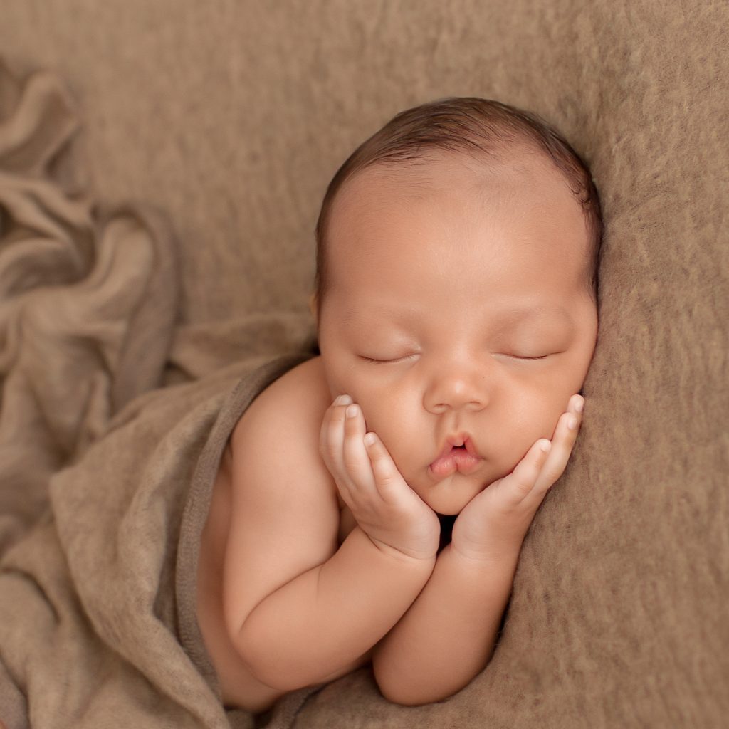 Lucas on brown blanket with baby hands cupping his beautiful baby face toward camera