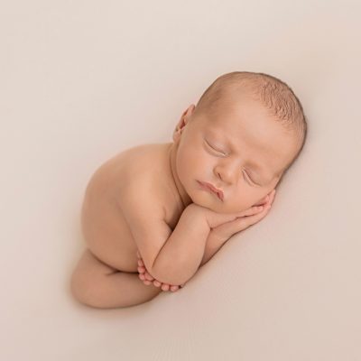 naked newborn baby sleeping with hands under cheeks and all ten teeny tiny toes wrapped around one little elbow against white blanket