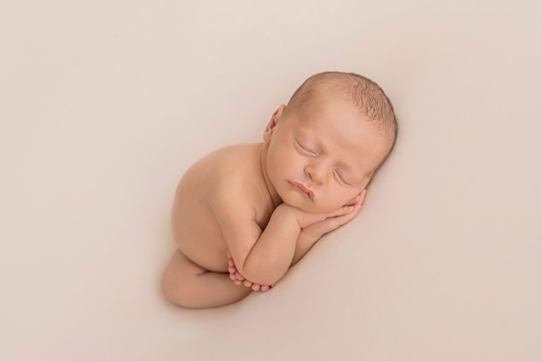 naked newborn baby sleeping with hands under cheeks and all ten teeny tiny toes wrapped around one little elbow against white blanket