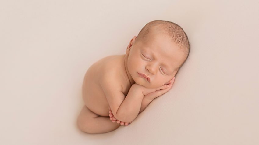 naked newborn baby sleeping with hands under cheeks and all ten teeny tiny toes wrapped around one little elbow against white blanket