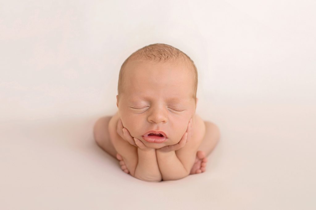naked newborn baby in froggy pose with hands cupping cheeks and chin and teeny tiny toes around his elbows against white blanket
