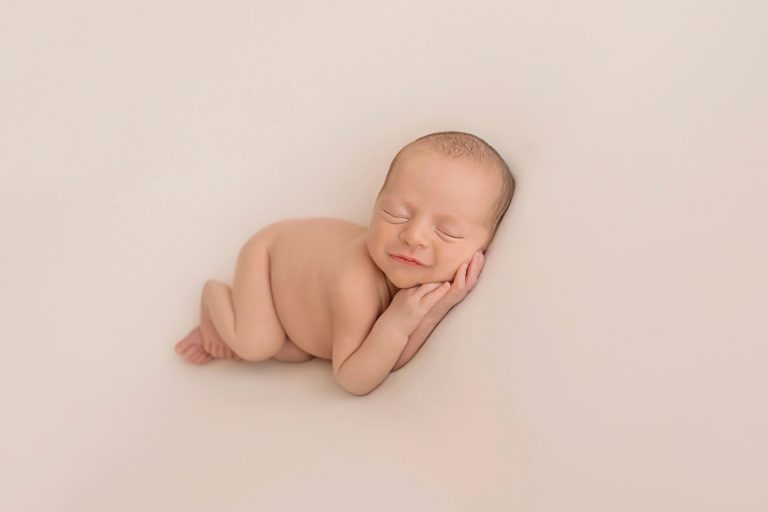 naked newborn baby smiling sleeping on side with tiny hands under cheeks against white blanket