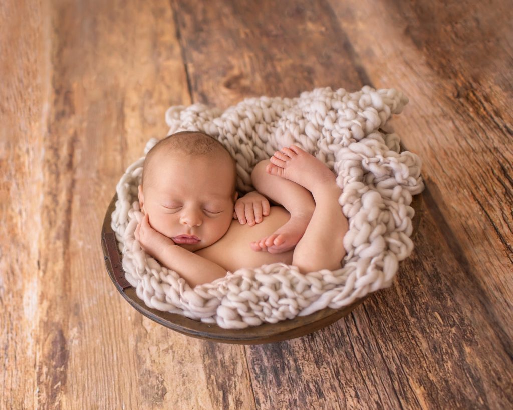 naked baby on beige chunky knit blanket in rustic brown bowl with against brown wood floor