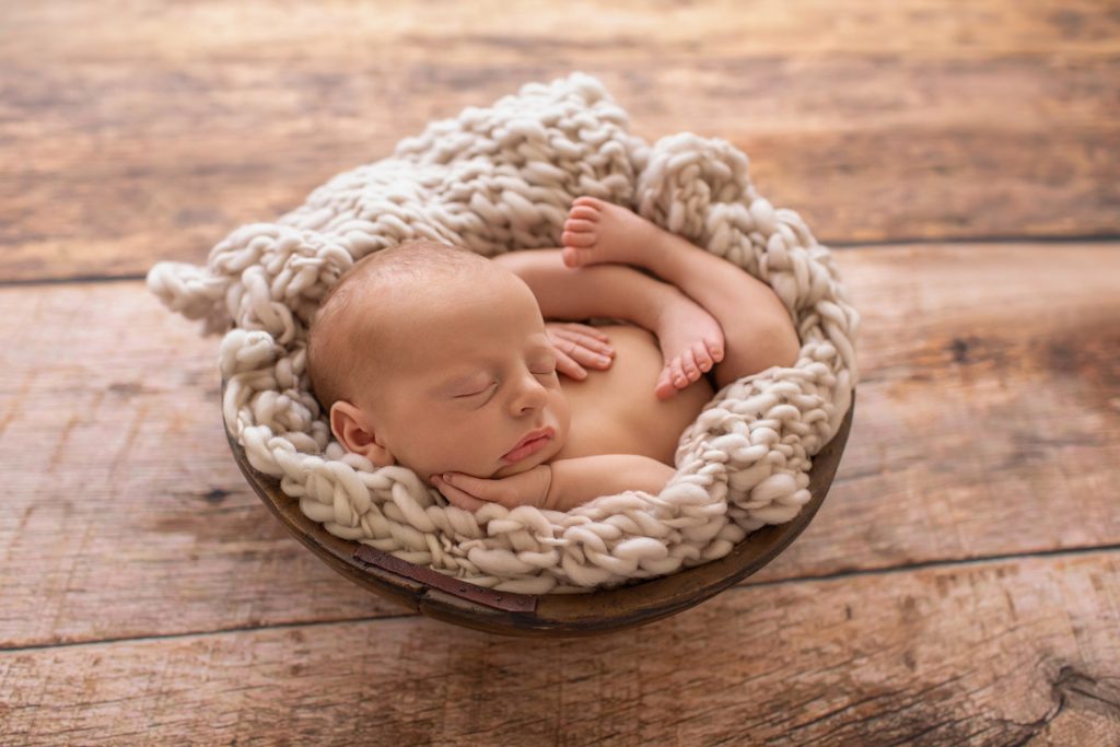 naked newborn baby on beige chunky knit blanket in rustic brown bowl against brown wood floor and backlight