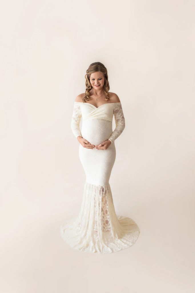 Pregnant mom long blond curls dressed in long sleeve ivory lace mermaid maternity gown looking down at beautiful baby belly