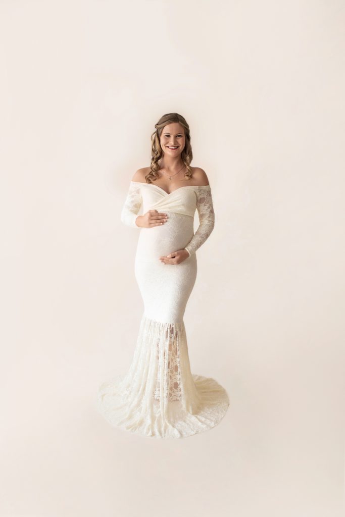 Pregnant mom curly blond hair dressed in long sleeve ivory lace mermaid maternity gown smiling at camera holding her beautiful baby belly