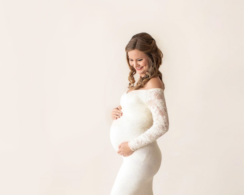 Pregnant mom long blond curl dressed in long sleeve ivory lace mermaid maternity gown profile smiling and looking at her beautiful baby belly