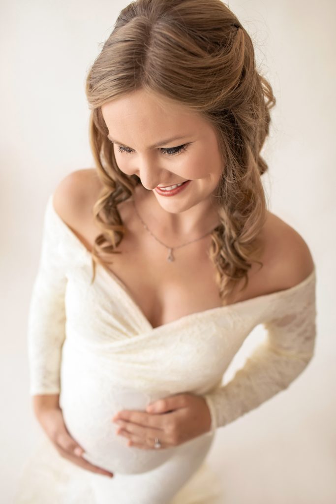 Pregnant mom curly blond hair dressed in long sleeve ivory lace mermaid maternity gown photo from above smiling and looking down at her beautiful baby belly