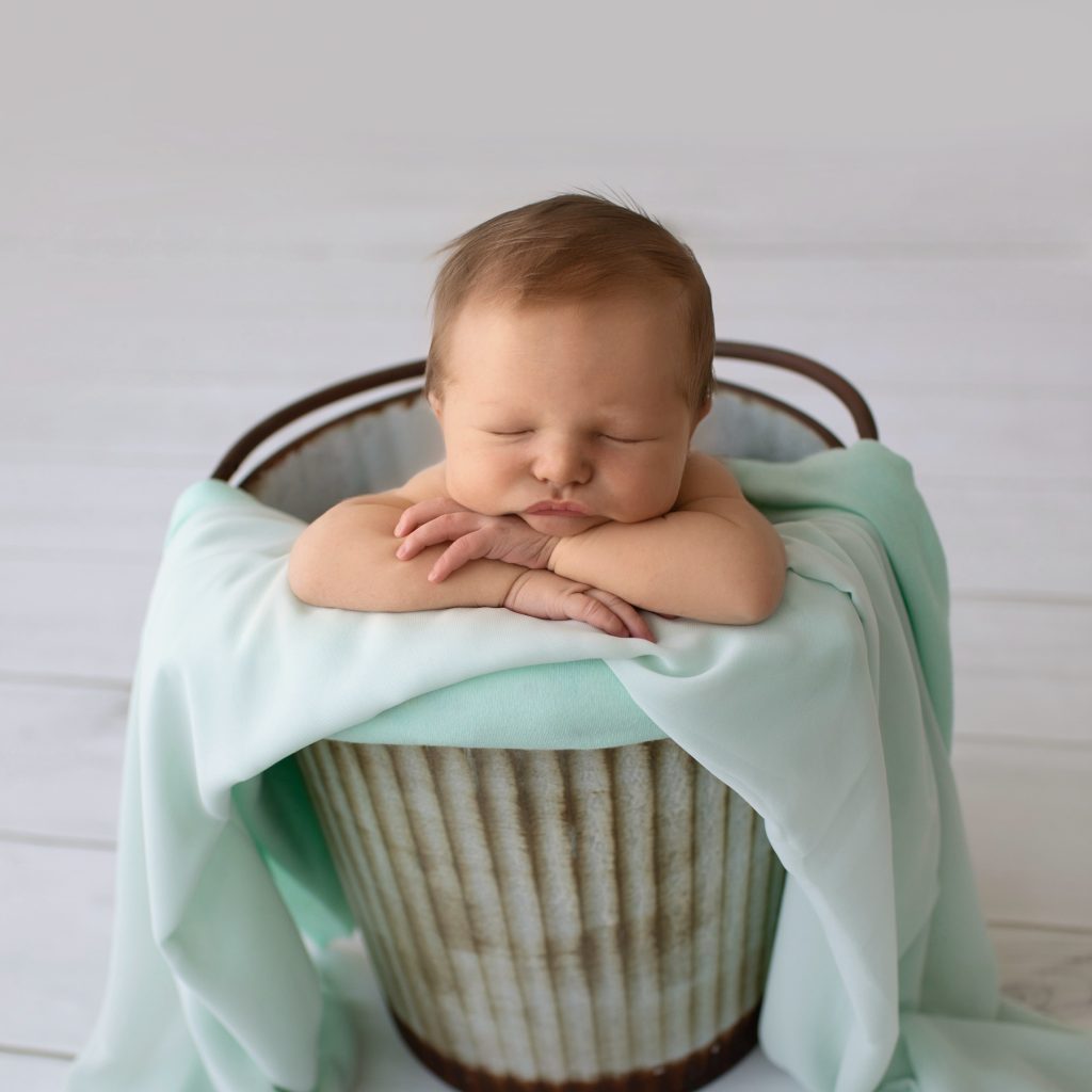 Newborn photography baby boy poses in metal bucket with mint green blanket on grey floor