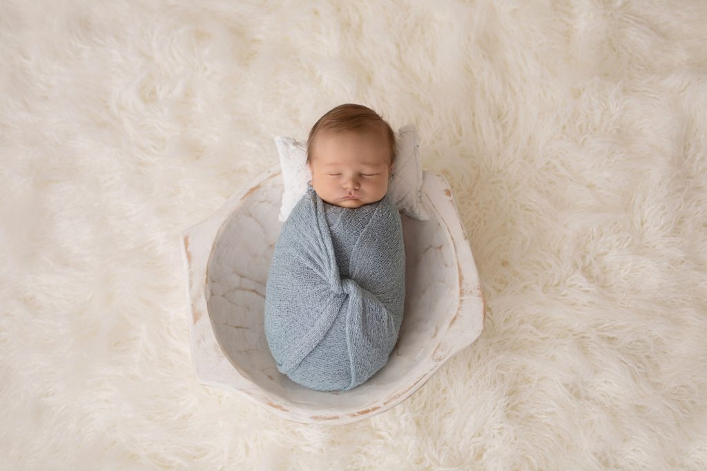 Newborn photography baby boy swaddled in pale blue posed in white wooden bowl on white fur