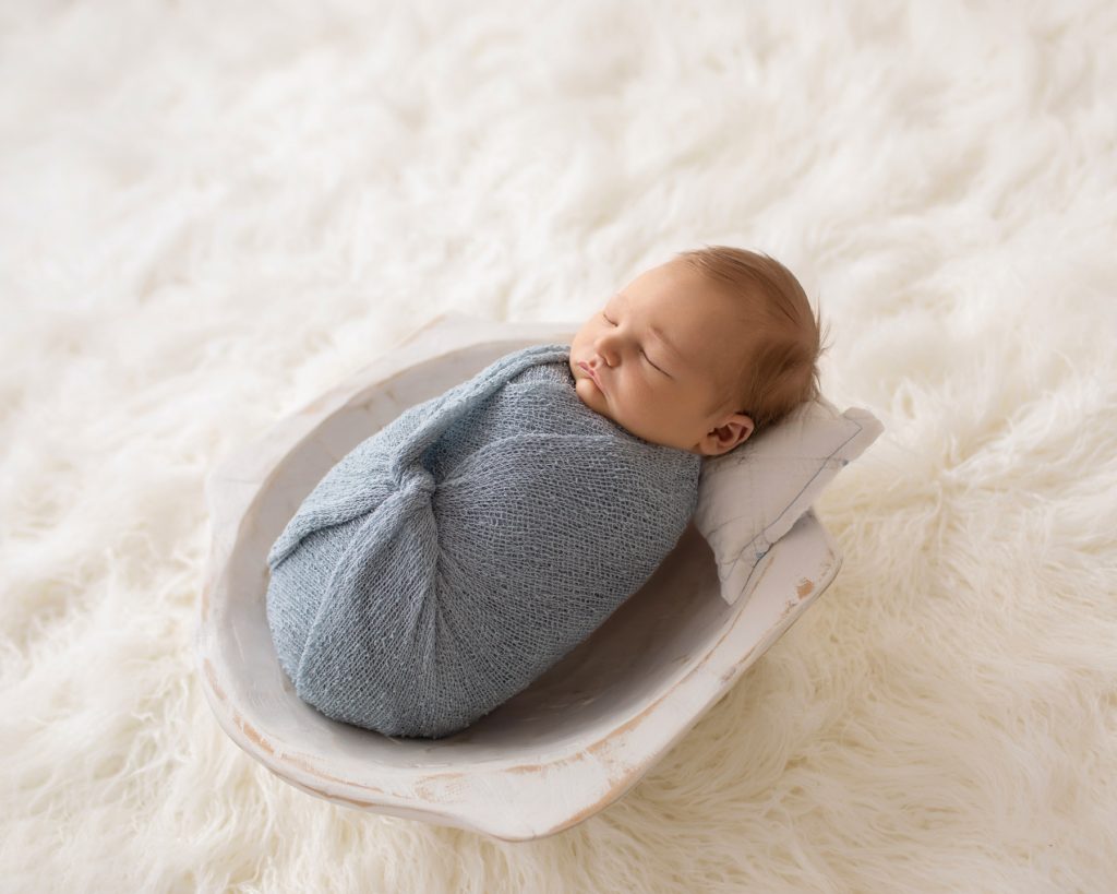 Newborn photography baby boy swaddled in pale blue posed in white wooden bowl on white fur with backlight