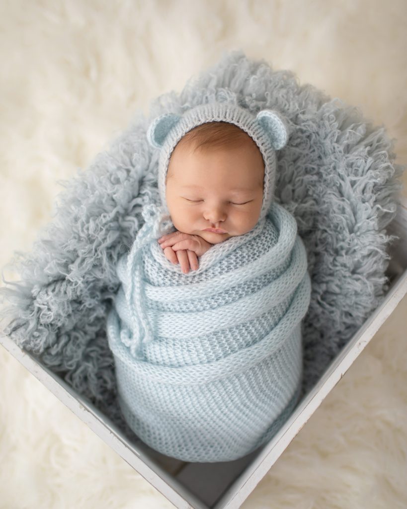 Newborn Gabe swaddled in a pale blue knit potato sack with matching bear bonnet in a fur stuffed white crate