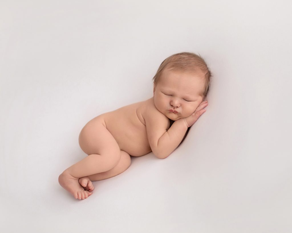Naked baby soft baby skin lounges on his side legs and toes stacked together and baby face resting on his hands
