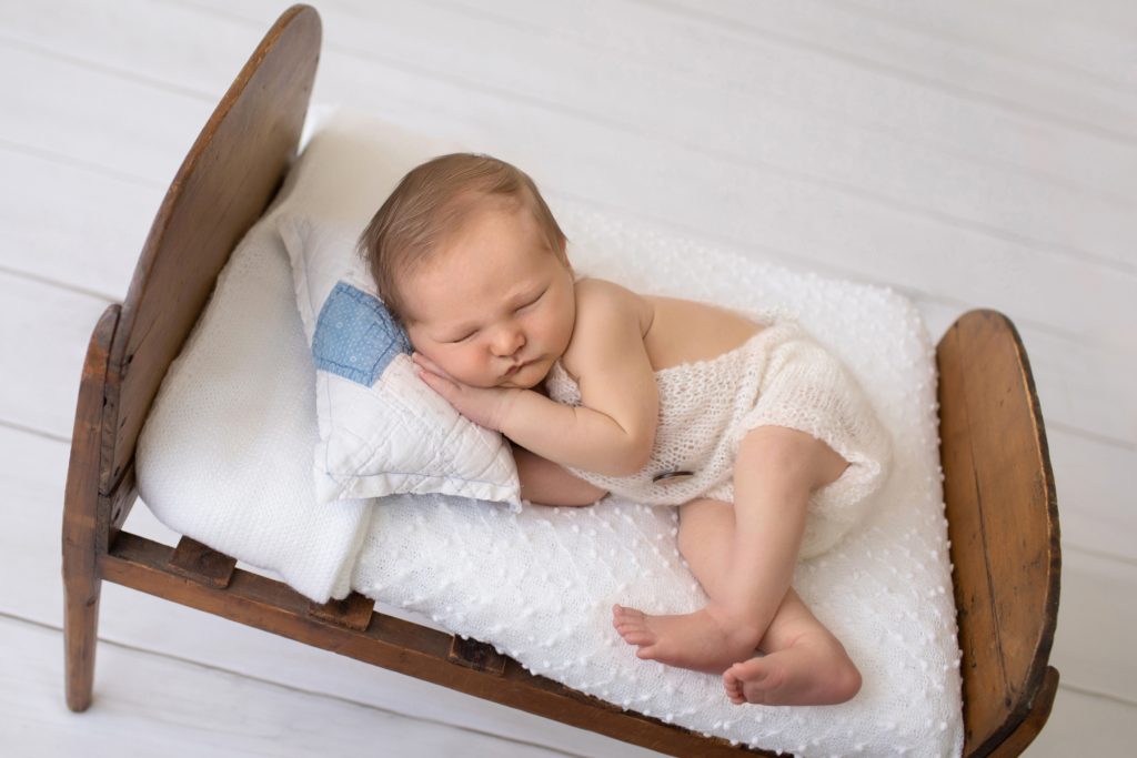 Newborn photography baby boy in knitted cream romper sleeps on wood bed baby prop face resting on hand feet crossed