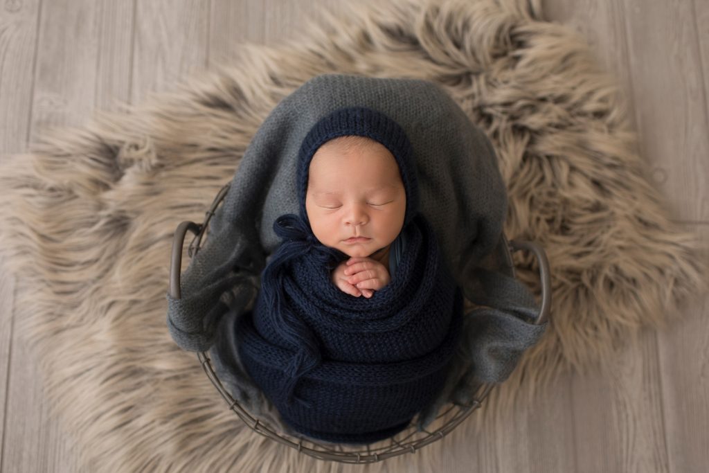 baby boy in navy knit blanket wrapped like potato sack with hands folded below chin matching navy knit bonnet posed in grey stuffed metal egg basket facing forward on grey wood floor