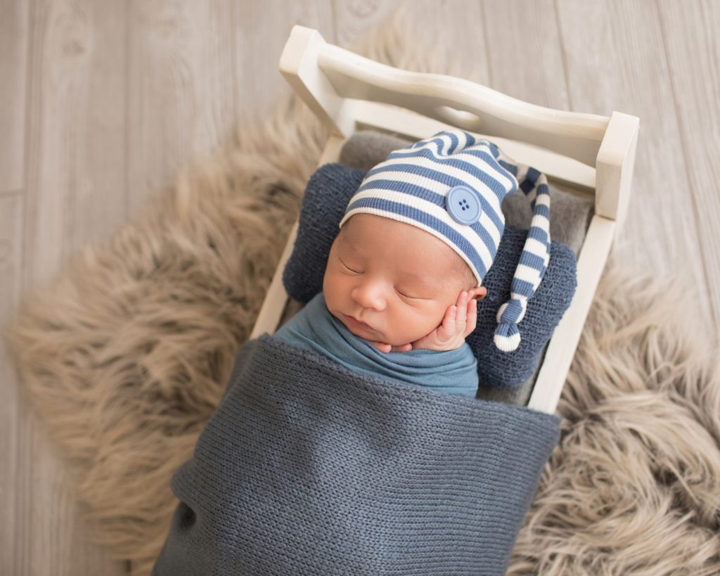 Newborn photography baby boy swaddled in blue with striped sleepy hat cupping his baby cheek with his tiny newborn hands sleeps on white wood bed baby prop Gainesville FL newborn mini photo session