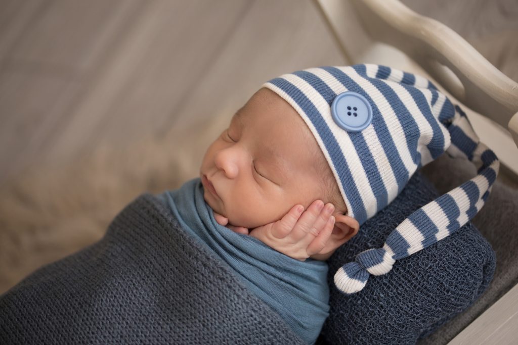Newborn photography baby boy swaddled in blue with striped sleepy hat cupping his baby cheek with his tiny newborn hands side view sleeps on white wood bed baby prop