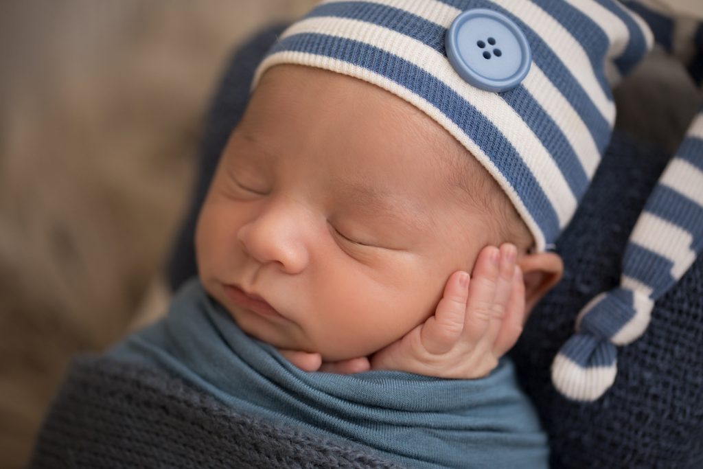 Newborn photography close up baby boy swaddled in blue with striped sleepy hat cupping his baby cheek with his tiny newborn hands sleeps on white wood bed baby prop