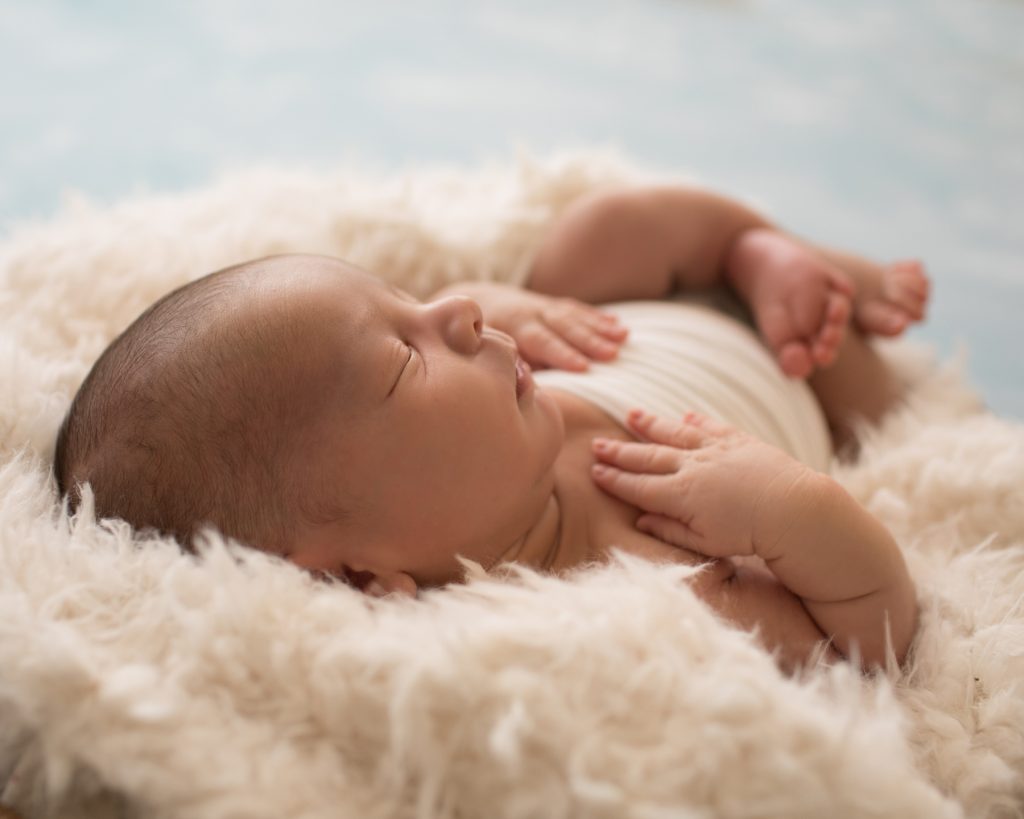 Newborn photography back lit baby boy in white swaddle sleeps in white fur stuffed wooden bowl against blue clouds