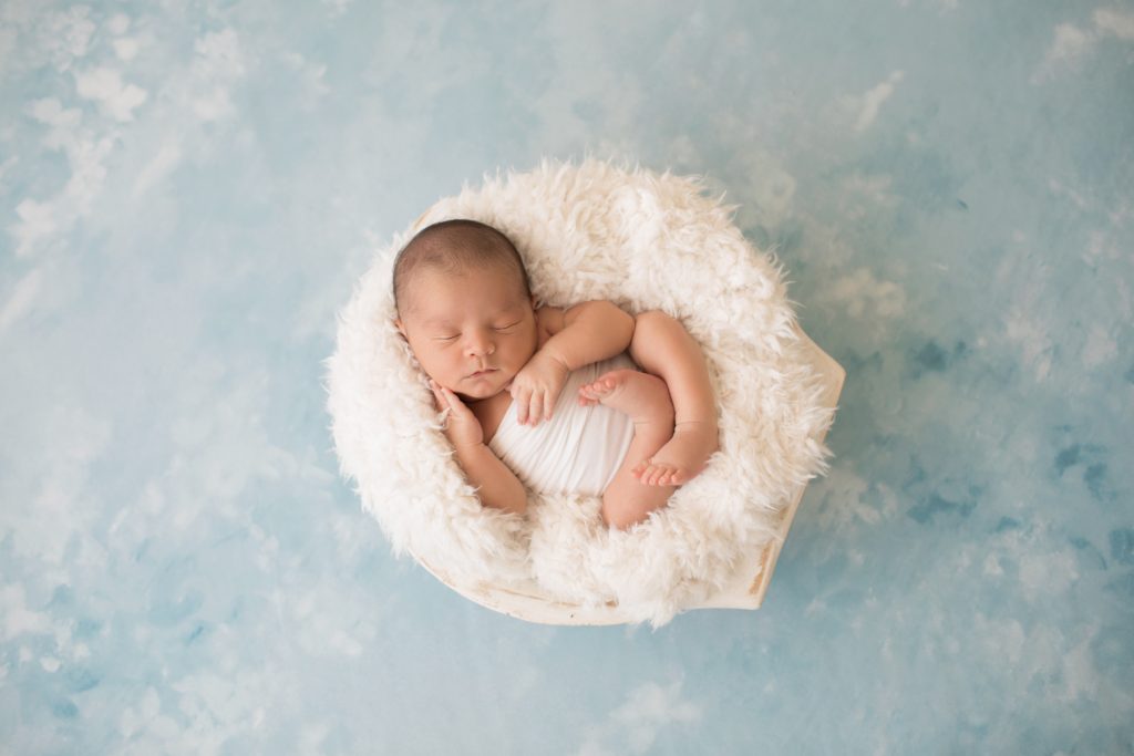 Newborn photography baby boy in white swaddle cupping his baby cheek with his tiny newborn hands sleeps in white fur stuffed wooden bowl on pale blue clouds Gainesville FL newborn mini photo session