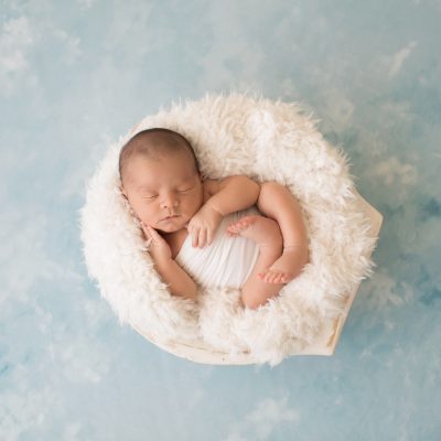 Newborn photography baby boy in white swaddle cupping his baby cheek with his tiny newborn hands sleeps in white fur stuffed wooden bowl on pale blue clouds Gainesville FL newborn mini photo session