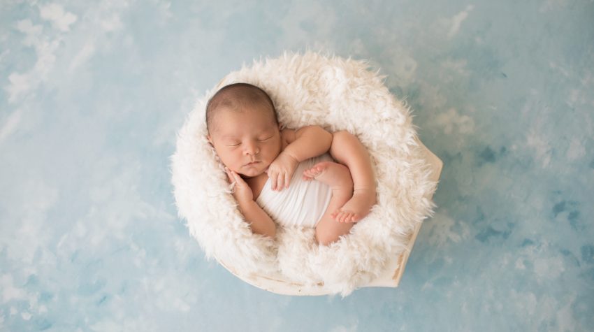 Newborn photography baby boy in white swaddle cupping his baby cheek with his tiny newborn hands sleeps in white fur stuffed wooden bowl on pale blue clouds Gainesville FL newborn mini photo session